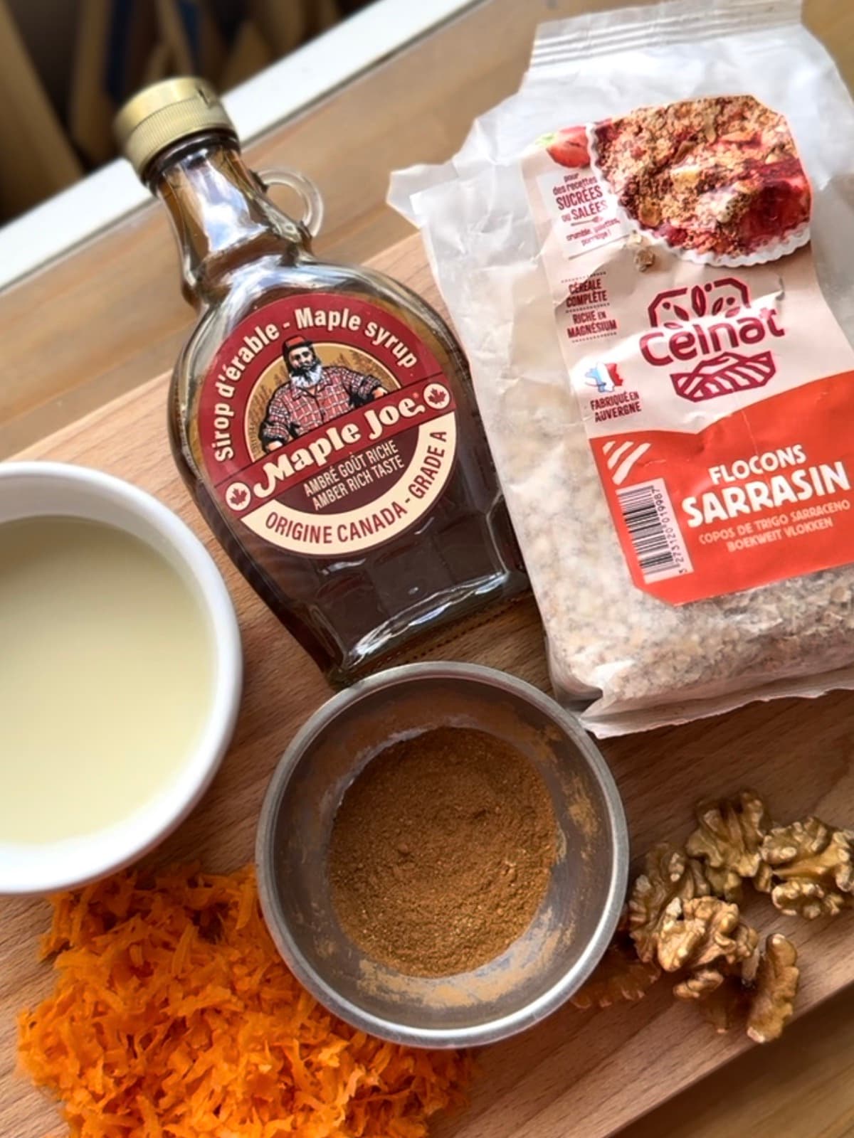 Fiocchi di grano saraceno, sciroppo d'acero, latte senza lattosio, carota grattugiata, cannella in polvere e noci su un tagliere di legno