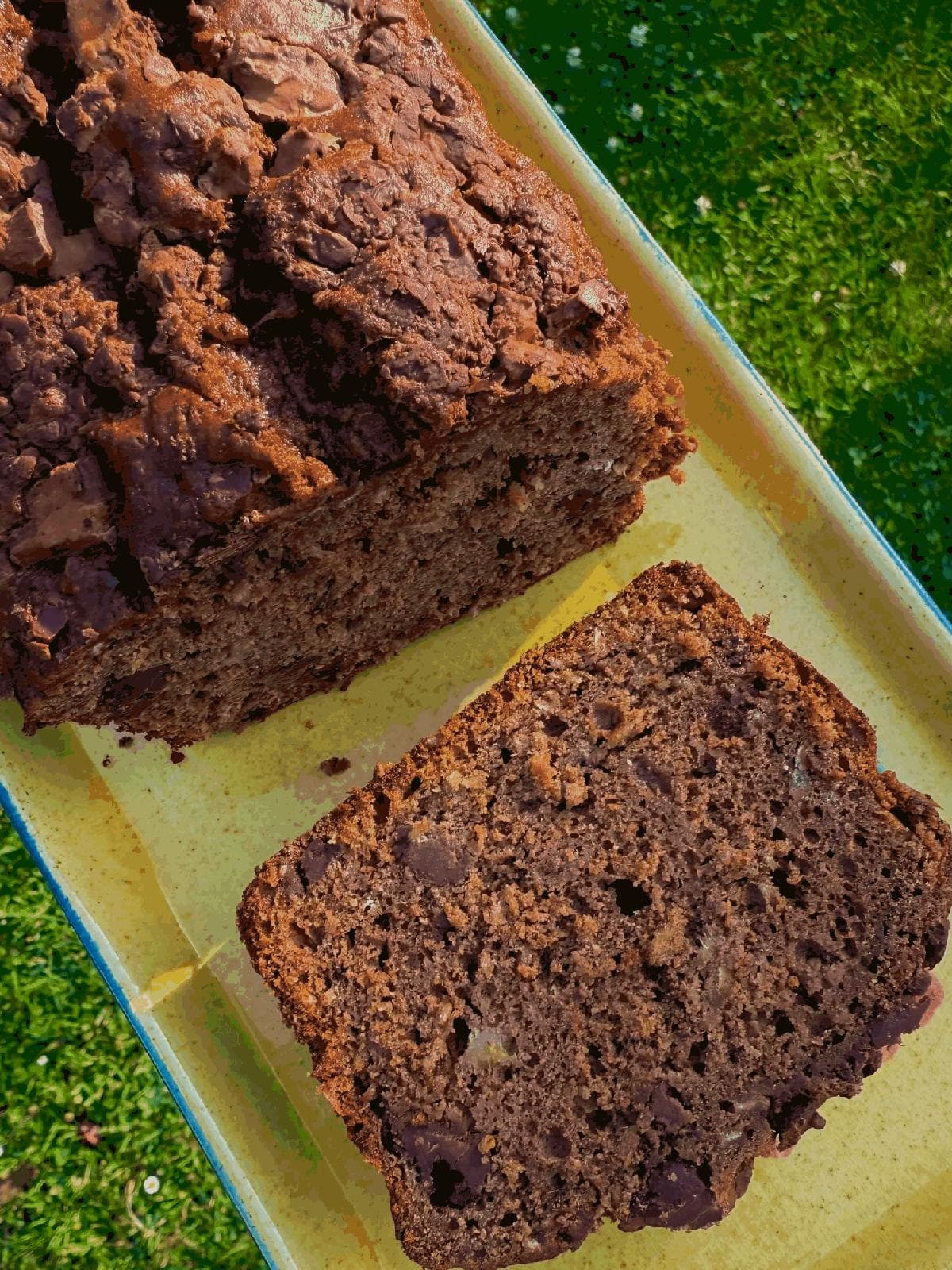 A chocolate banana bread loaf with a thick slice cut off, on a yellow dish on the grass